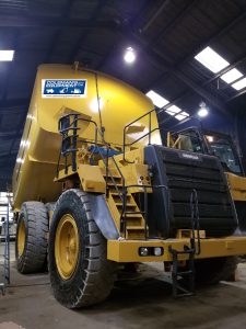 A large yellow dump truck inside an industrial building. The truck has a raised platform with a staircase leading to the cabin, and overhead lights illuminate the space.