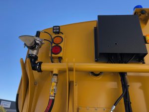 Close-up of the back of a yellow utility vehicle, showing a red and amber light, various pipes, wires, and a black box. Bright blue sky in the background.